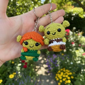 Puede incluir: Dos llaveros hechos a mano de ganchillo con personajes de dibujos animados. Uno tiene pelo naranja y un atuendo verde, el otro tiene un cuerpo verde, un atuendo marrón y blanco. Ambos tienen ojos negros y están unidos a llaveros plateados.