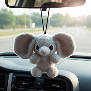 May include: A gray plush elephant car charm hanging from a rearview mirror. The elephant has large ears, a small trunk, and black button eyes. The charm is suspended by a black string, adding a touch of whimsy to the car's interior.