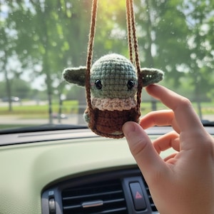 May include: A handmade crochet Baby Yoda car charm. The figure is green with large black eyes, tan accents, and a brown base, suspended by brown cords. The charm is hanging in front of a car dashboard.