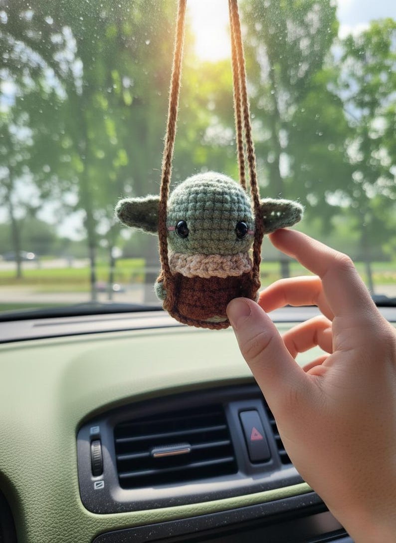 May include: A handmade crochet Baby Yoda car charm. The figure is green with large black eyes, tan accents, and a brown base, suspended by brown cords. The charm is hanging in front of a car dashboard.