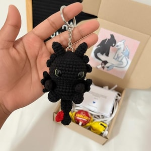 以下が含まれることがあります： 緑色の目と赤い尾を持つ、黒いかぎ針編みのドラゴンのキーホルダー。 ドラゴンはシルバーのキーリングに取り付けられています。 キーホルダーは、箱、カード、お菓子を含むギフトセットの一部です。
