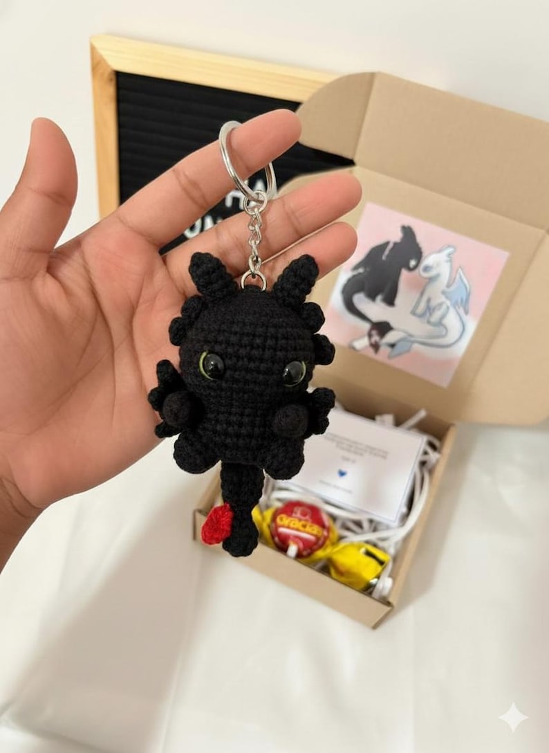 以下が含まれることがあります： 緑色の目と赤い尾を持つ、黒いかぎ針編みのドラゴンのキーホルダー。 ドラゴンはシルバーのキーリングに取り付けられています。 キーホルダーは、箱、カード、お菓子を含むギフトセットの一部です。