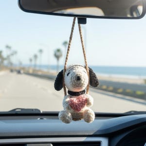 May include: A crocheted Snoopy car charm, hanging from the rearview mirror. The charm features a white body, black ears, and a pink heart. The background shows a road and the ocean.