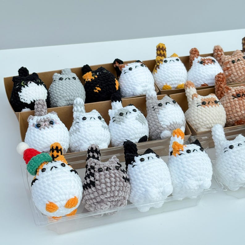 Puede incluir: Una colecci&oacute;n de figuras de gato de ganchillo hechas a mano en varios colores y patrones. Los gatos se exhiben en una bandeja de pl&aacute;stico transparente y contenedores de cart&oacute;n. Algunos gatos tienen marcas negras, grises, naranjas y blancas. Un gato lleva un peque&ntilde;o gorro de Navidad.