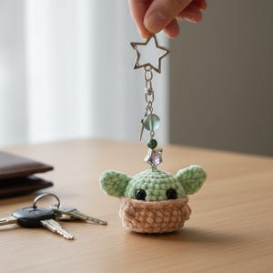 May include: Crocheted keyring featuring a green and beige character with black eyes and large ears. The keyring has a star-shaped clasp and decorative beads. The character is in a beige basket-like container.
