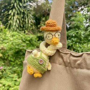 Puede incluir: Un pato hecho a mano con ganchillo que lleva un sombrero marrón y gafas, un chaleco verde y una bufanda amarilla. El pato está sujeto a un bolso tote beige. El fondo es una vista borrosa de follaje verde.