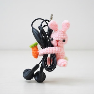 May include: A handmade pink crochet bunny holding a small orange and green carrot, wrapped around a pair of black earbuds and their cord. The bunny has black eyes and a red nose. The background is a plain white.