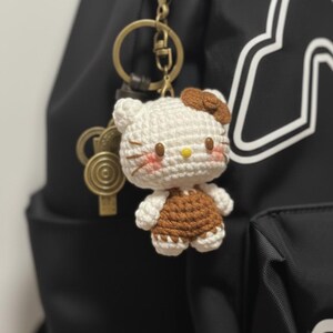May include: A handmade crochet Hello Kitty keychain. The white and brown figure has a yellow nose, rosy cheeks, and a brown bow. The keychain is attached to a gold-colored clasp and a black backpack.