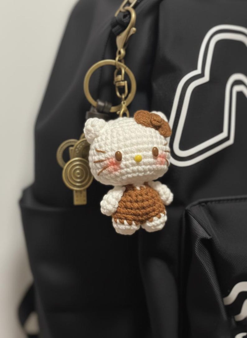 May include: A handmade crochet Hello Kitty keychain. The white and brown figure has a yellow nose, rosy cheeks, and a brown bow. The keychain is attached to a gold-colored clasp and a black backpack.