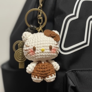 May include: A handmade crochet keychain featuring a white and brown Hello Kitty design. The keychain has a gold-colored clasp and is attached to a black backpack. The Hello Kitty figure has a brown bow, rosy cheeks, and a yellow nose.