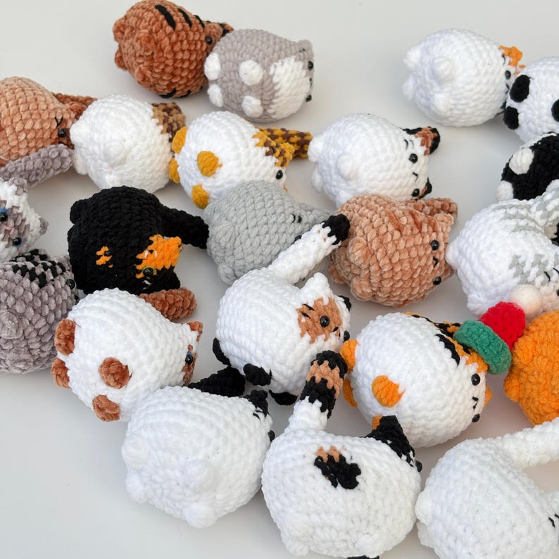 Puede incluir: Una colecci&oacute;n de juguetes de gato hechos a mano con ganchillo en varios colores y patrones. Los juguetes presentan cuerpos blancos, marrones, negros y grises con rasgos faciales y marcas detalladas. Algunos gatos tienen patrones rayados o manchados, mientras que otros son de color s&oacute;lido. Algunos tienen sombreros peque&ntilde;os.