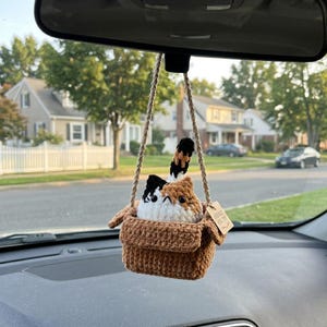 May include: A crocheted calico cat ornament sitting inside a brown crocheted box. The cat has white, orange, and black yarn colors with black eyes and a small tail pointing upward. It hangs from a braided jute cord and includes a small tan tag.