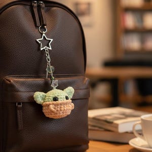 May include: A brown leather backpack with a crocheted charm. The charm is a light green and tan character with a star-shaped metal charm attached to the zipper. The background is blurred, showing a table and a bookshelf.