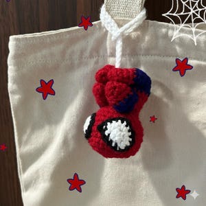 Könnte beinhalten: Ein roter, blauer und weißer gehäkelter Spider-Man-Anhänger, der an einer beigen Tragetasche hängt. Der Anhänger zeigt ein detailliertes Design von Spider-Mans Maske und Hand. Die Tasche ist mit roten sternförmigen Aufklebern verziert.