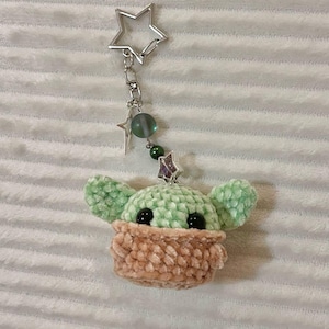 May include: A handmade keychain featuring a crocheted figure with large ears and a tan-coloured face covering. The keychain has a silver star-shaped clasp and decorative beads. The figure is light green and tan.