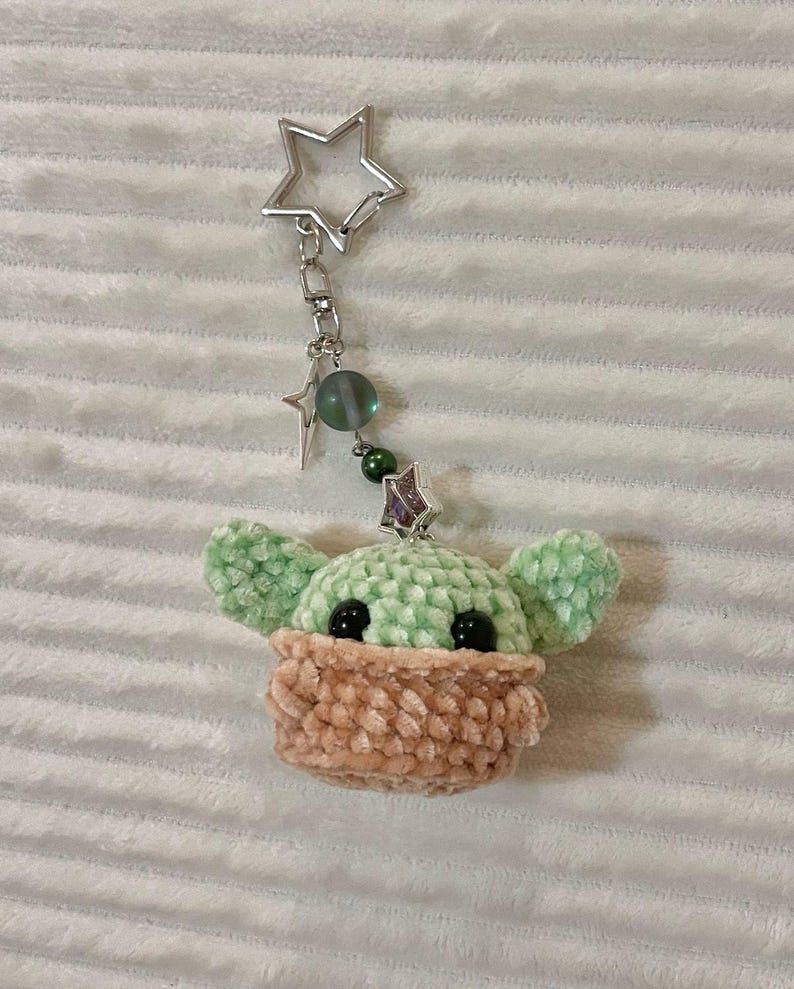 May include: A handmade keychain featuring a crocheted figure with large ears and a tan-coloured face covering. The keychain has a silver star-shaped clasp and decorative beads. The figure is light green and tan.