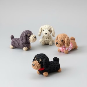 May include: Four crocheted dachshund dog figurines in various colours: purple, cream, brown, and black. The brown dog wears a pink and white sweater, and the black dog has a red collar. Each dog has a unique expression and pose.