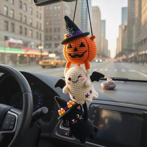 Puede incluir: Un adorno de ganchillo colgante que presenta una pila de tres figuras: una calabaza naranja con un sombrero de bruja negro con diseño de telaraña, un fantasma blanco con alas negras y estrellas moradas, y un gato negro con sombrero y dulces.