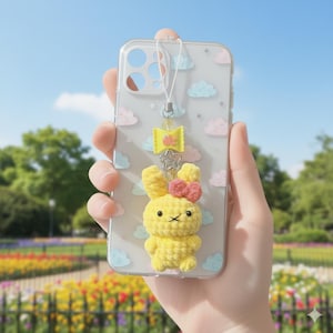 Puede incluir: Funda transparente para teléfono con un dije de conejo de ganchillo amarillo, un lazo rosa y un dije en forma de lazo amarillo. La funda tiene un patrón de nubes con nubes rosas y azules. Una correa blanca está unida a la funda.