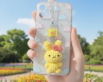 May include: Clear phone case with a yellow crochet bunny charm, a pink bow, and a yellow bow-shaped charm. The case has a cloud pattern with pink and blue clouds. A white lanyard is attached to the case.