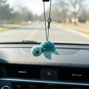 May include: A crocheted turtle car charm in shades of teal and turquoise, hanging from a black string. The turtle has a rounded head, a textured shell, and small flipper-like appendages. The charm is suspended in front of a car dashboard.