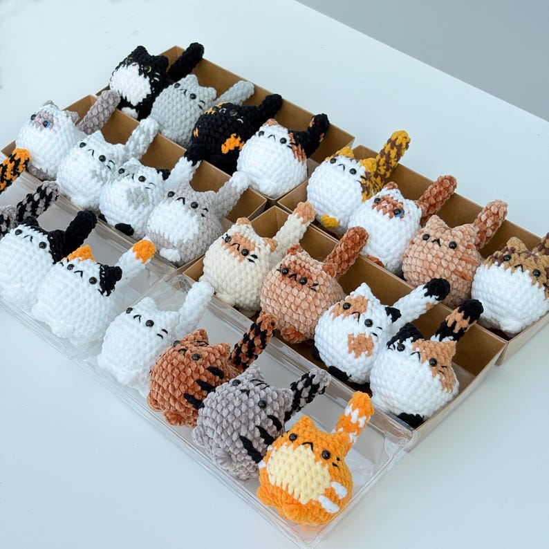 Puede incluir: Una colecci&oacute;n de figuras de gato de ganchillo hechas a mano en varios colores, incluyendo blanco, negro, naranja y gris. Cada gato tiene un patr&oacute;n &uacute;nico y est&aacute; dispuesto en compartimentos individuales dentro de una bandeja de pl&aacute;stico transparente.