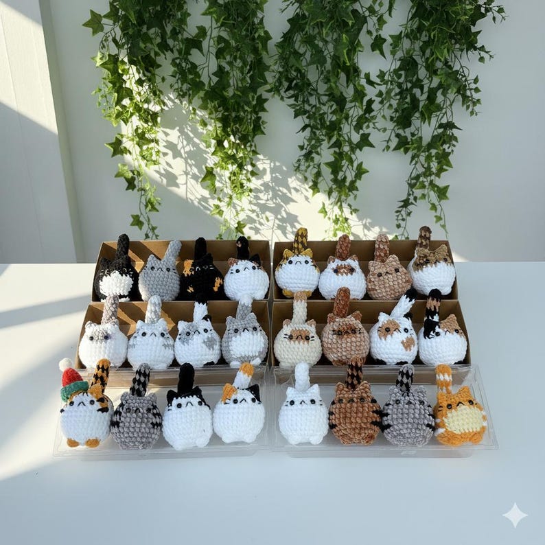Puede incluir: Una colecci&oacute;n de figuras de gatos de ganchillo hechas a mano en varios colores y patrones. Los gatos se exhiben en un estante escalonado, con una variedad de colores que incluyen blanco, negro, gris, marr&oacute;n y naranja. El fondo presenta plantas verdes colgantes.