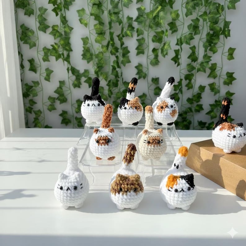 Puede incluir: Una colecci&oacute;n de figuras de gato de ganchillo hechas a mano en varios colores y patrones. Los gatos son blancos con marcas negras, marrones y naranjas. Cada gato tiene una cola hacia arriba. Las figuras se exhiben en un soporte transparente de varios niveles.