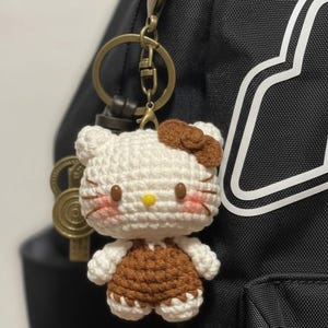 May include: A handmade crochet keychain featuring a white and brown Hello Kitty design. The keychain has a gold-colored clasp and key ring. The figure has a brown bow, pink cheeks, and a yellow nose. The keychain is attached to a black backpack.