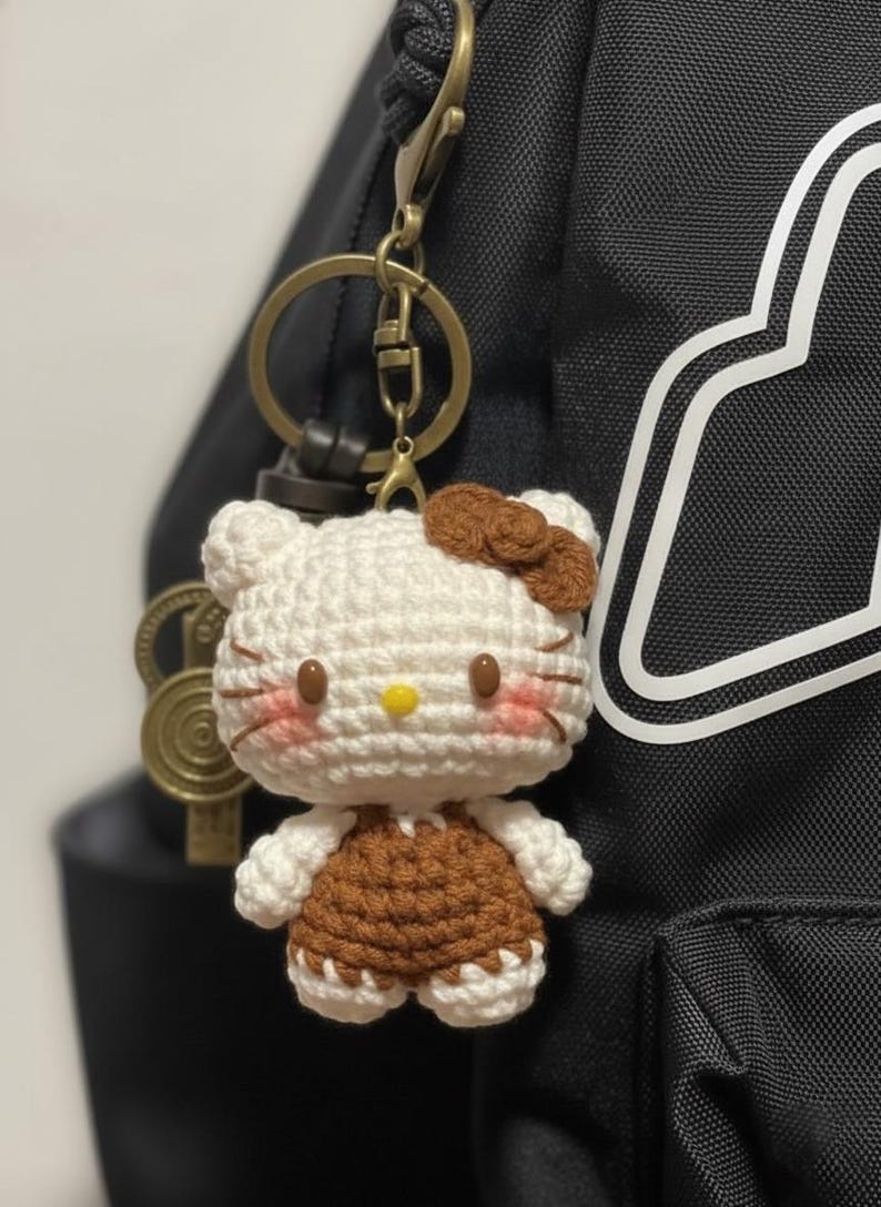May include: A handmade crochet keychain featuring a white and brown Hello Kitty design. The keychain has a gold-colored clasp and key ring. The figure has a brown bow, pink cheeks, and a yellow nose. The keychain is attached to a black backpack.
