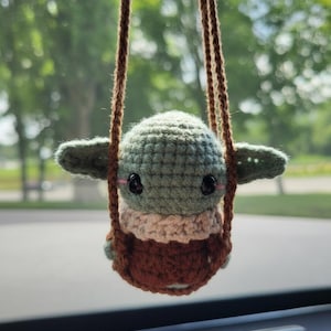 May include: A crocheted Baby Yoda doll in a brown swing. The doll has large black eyes, green skin, and a tan and brown outfit. The swing is suspended by brown cords, with a blurred green background.