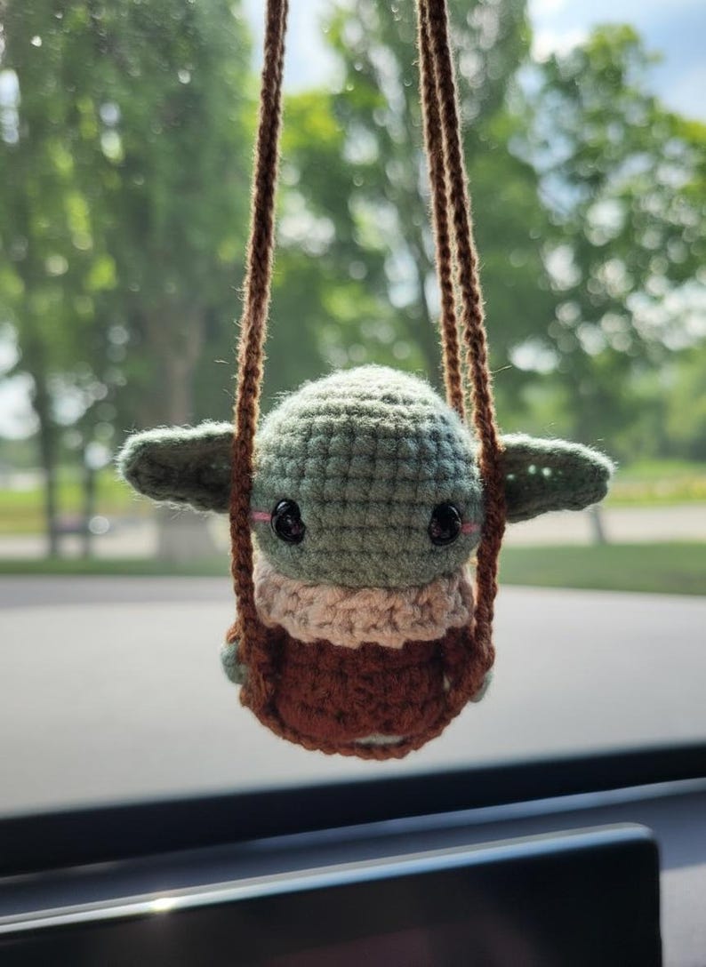 May include: A crocheted Baby Yoda doll in a brown swing. The doll has large black eyes, green skin, and a tan and brown outfit. The swing is suspended by brown cords, with a blurred green background.