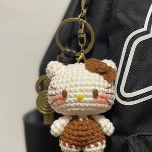 May include: A handmade crochet keychain featuring a Hello Kitty design. The keychain has a white head with brown accents, including a bow and overalls. The face has pink cheeks, a yellow nose, and brown whiskers. The keychain is attached to a gold-colored metal ring.