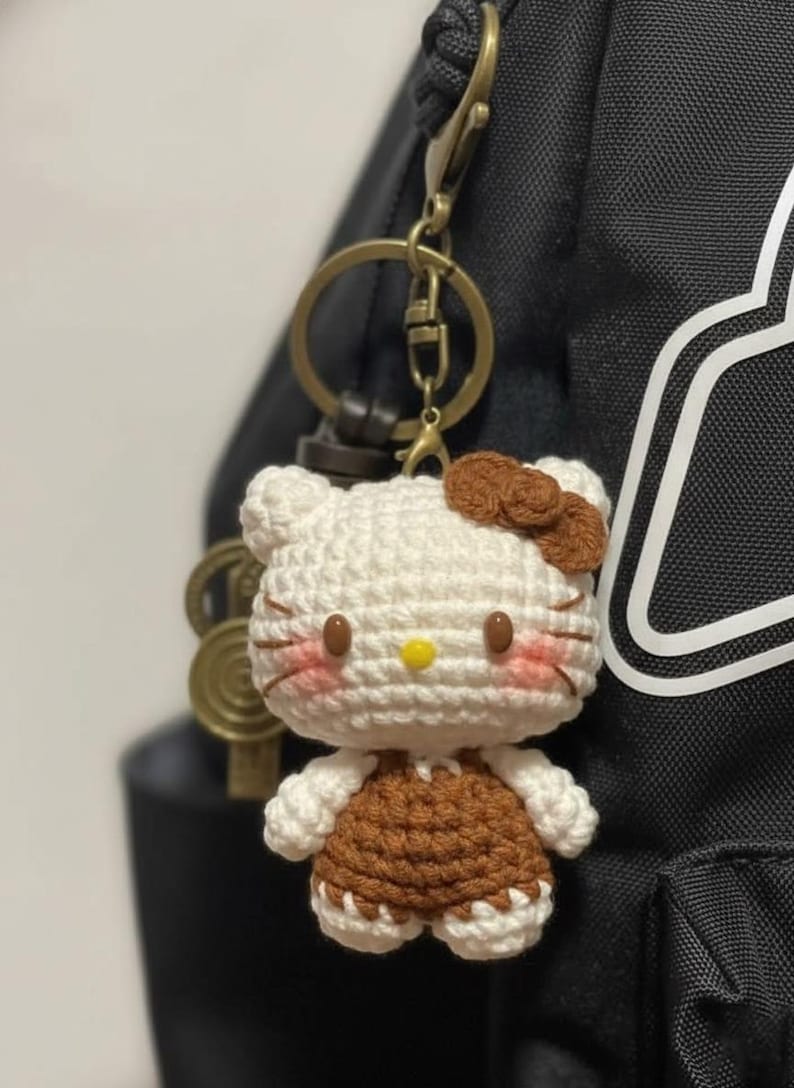 May include: A handmade crochet keychain featuring a Hello Kitty design. The keychain has a white head with brown accents, including a bow and overalls. The face has pink cheeks, a yellow nose, and brown whiskers. The keychain is attached to a gold-colored metal ring.