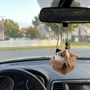 Pu&ograve; includere: Un peluche a forma di gatto calico all'uncinetto seduto dentro una scatola di filato marrone, appeso a uno specchietto retrovisore con uno spago di juta. Un piccolo cartellino rettangolare riporta il testo stampato, HANDMADE WITH LOVE.