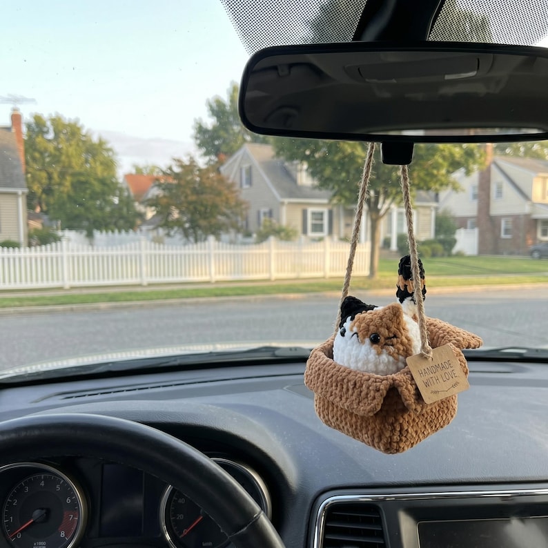 Pu&ograve; includere: Un peluche a forma di gatto calico all'uncinetto seduto dentro una scatola di filato marrone, appeso a uno specchietto retrovisore con uno spago di juta. Un piccolo cartellino rettangolare riporta il testo stampato, HANDMADE WITH LOVE.