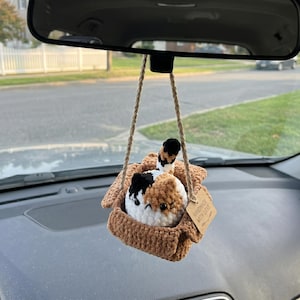 Pu&ograve; includere: Un gatto di peluche tricolore all'uncinetto seduto in una piccola scatola marrone all'uncinetto, appeso a un cordino beige. Il gatto presenta filati bianchi, marroni e neri con occhi a perla neri. Un'etichetta di carta riporta HANDMADE WITH LOVE.
