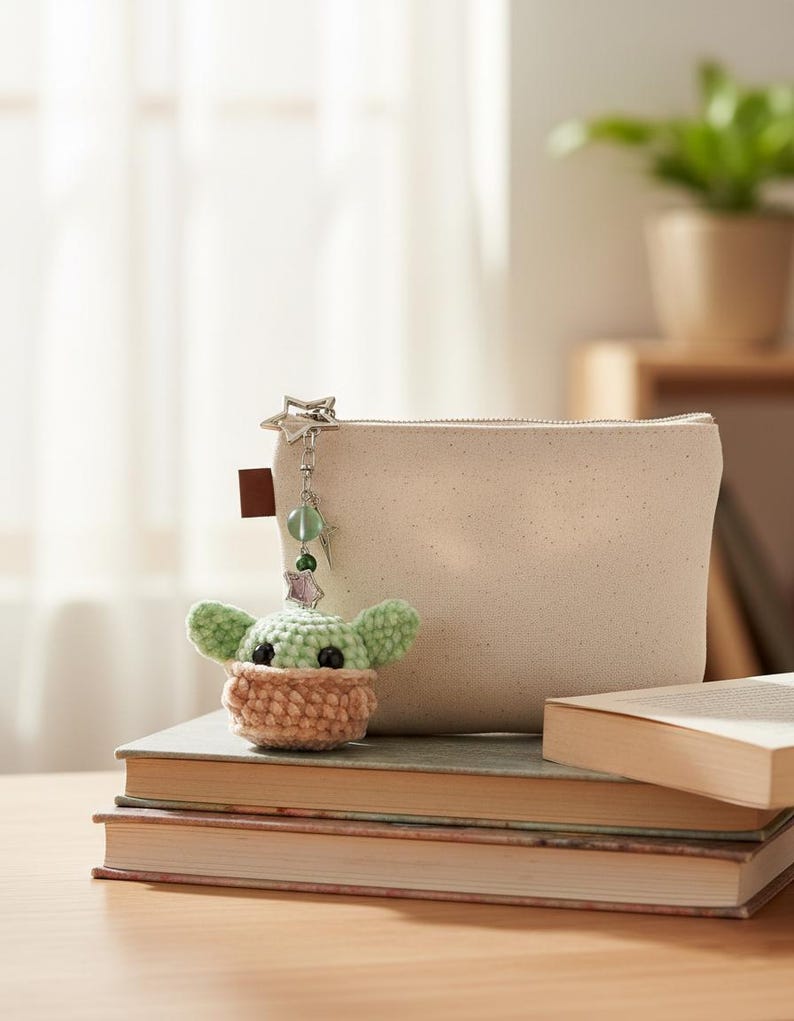 May include: A crocheted character charm with large ears and a tan muzzle, suspended from a silver keychain with a star charm. It sits beside a beige canvas pouch and a stack of books.