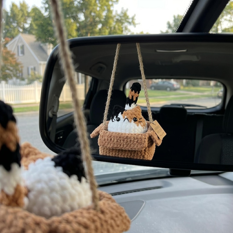 Pu&ograve; includere: Un peluche di gatto calico all'uncinetto seduto dentro una scatola marrone all'uncinetto, sospeso da uno spago di juta. Il gatto ha macchie di lana bianca, nera e arancione, occhi di sicurezza neri e un cartellino marrone sul lato.
