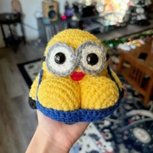 May include: A handmade, crocheted Minion character with yellow body, blue overalls, and large, round eyes. The Minion has a red mouth and is held in a hand. The background is blurred.