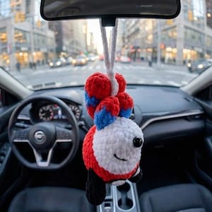 May include: A handmade car charm featuring a smiling fox design. The fox is crocheted with red, white, blue, and black yarn, hanging from a white cord. The charm is suspended from a car's rearview mirror, with a city street visible in the background.