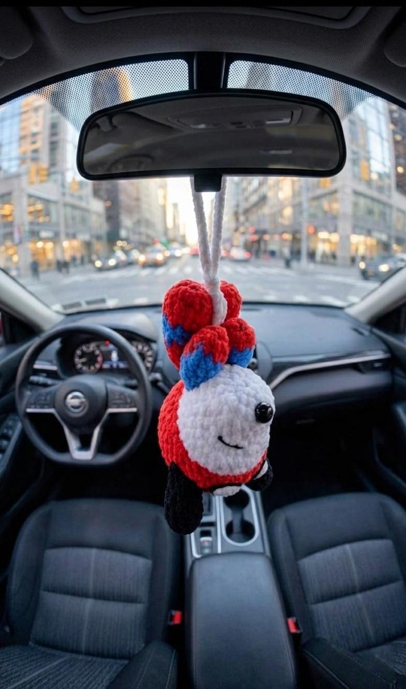 May include: A handmade car charm featuring a smiling fox design. The fox is crocheted with red, white, blue, and black yarn, hanging from a white cord. The charm is suspended from a car's rearview mirror, with a city street visible in the background.