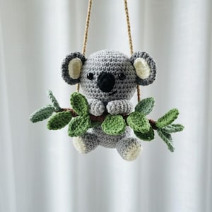 May include: A crocheted koala bear in grey and white, with a black nose and small black eyes. The koala holds a branch with green leaves and hangs from a brown rope. A delightful, handmade soft toy, perfect for a nursery.