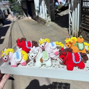 May include: A collection of handmade crochet horse figurines in various colors, including pink, red, white, and orange. Each horse features a unique design with yellow, blue, and green accents. The horses are displayed on a white tray.