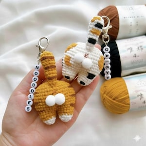 May include: Two handmade crochet cat butt keyrings. One is orange with white accents and the other is white with orange and black accents. Each keyring has a beaded name tag. The background includes spools of yarn.