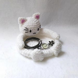 Könnte beinhalten: Ein weißes, gehäkeltes Katzen-Schmucktablett mit einem schwarzen Armband, einer silbernen Uhr und einem schwarzen, blütenförmigen Ring. Die Katze hat rosa Innenohren und schwarze Gesichtsdetails. Für kleinen Schmuck.