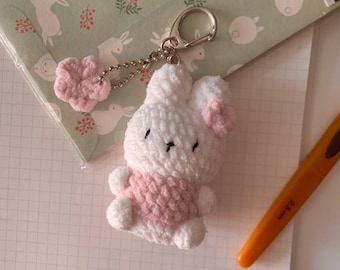 May include: A handmade white and pink crochet bunny keychain. The bunny has pink ears and a pink sweater. A pink flower charm hangs from the keychain. The background includes a green card with rabbit illustrations and a grid-lined paper.