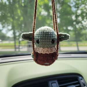 May include: A crocheted Baby Yoda car charm, suspended by a brown cord. The charm has a green head, large black eyes, and a brown and beige outfit. The background shows a car interior and a blurred view of trees.