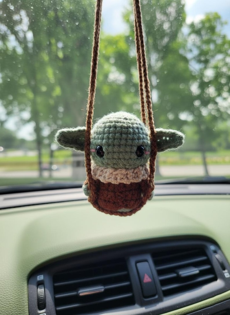 May include: A crocheted Baby Yoda car charm, suspended by a brown cord. The charm has a green head, large black eyes, and a brown and beige outfit. The background shows a car interior and a blurred view of trees.