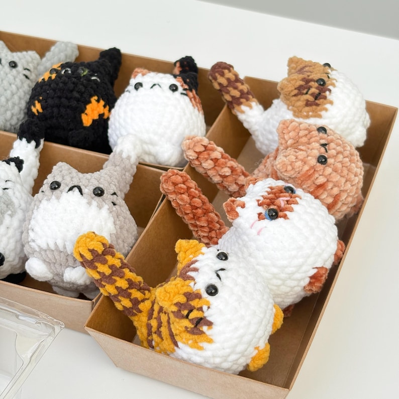 Puede incluir: Una colecci&oacute;n de juguetes de gato hechos a mano con ganchillo en una caja de cart&oacute;n. Los gatos son blancos, grises, negros y naranjas, con varios patrones. Cada gato tiene ojos de bot&oacute;n negros y una cola larga. Los juguetes son suaves y afelpados.